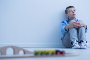 child sitting against a plain white wall, holding arms around legs and looking up at something. He looks sad and upset.