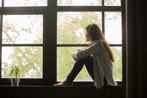 Depressed teen girl sits on a window sill and stares outside.