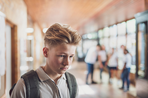 A boy walks alone down a school hallway with a worried look on his face while a group of kids laugh in the background.